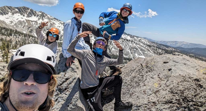 Five people wearing helmets stand atop a rocky summit overlooking snow capped mountains. One of them flexes their muscles. 